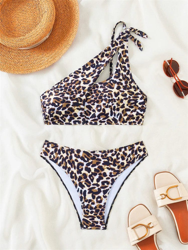Maillot de bain bikini asymétrique léopard avec fond beige accompagné de lunettes et chapeau.