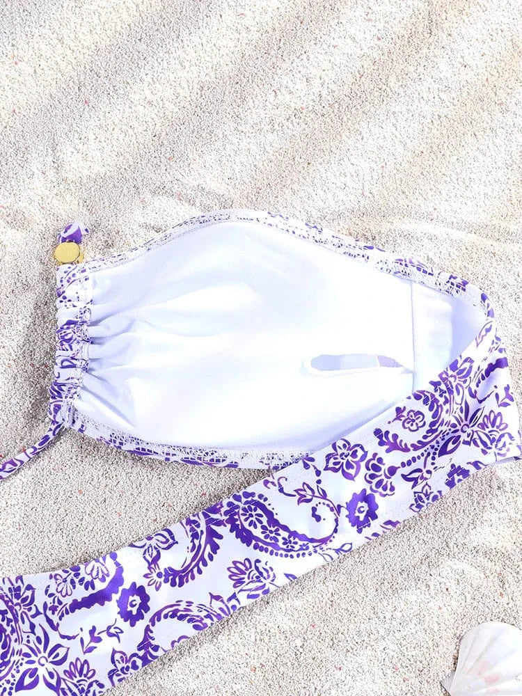 Maillot de bain femme avec motifs violets et blancs, style triangle ficelle, posé sur le sable.