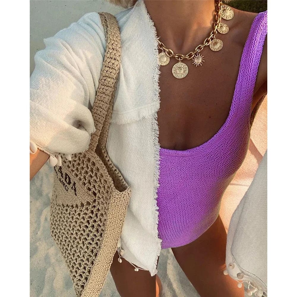 Maillot de bain femme 1 pièce violet, porté avec un sac en paille et un collier doré sur la plage.